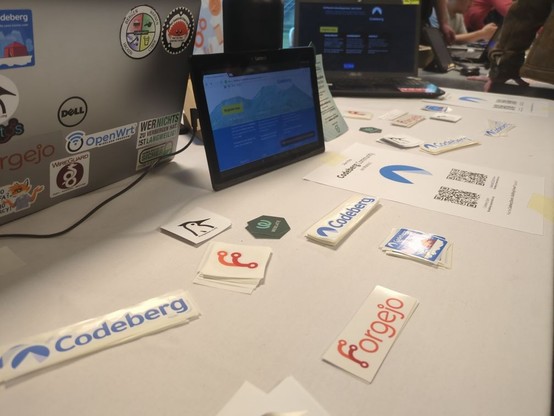 Close-up shot of stickers on our tabel at FOSDEM. Two laptops and a tablet present Codeberg's website and services.