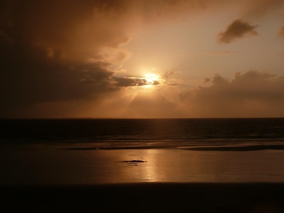 Abendsonne an leicht bewölktem Himmel über dem ruhigen Meer. Vorn im Bild Inselstrand