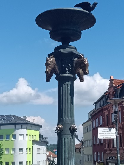 Das Bild zeigt den oberen Teil des Brunnens auf dem Synagogenplatz am Oberen Markt in Neunkirchen. Eine Bronzesäule mit Löwen- und Pferdeköpfen als Ornamente. Obenauf ist eine bronzene Schale mit einem auf dem Rand befindlichen Vogel (Bronze). 
Rechts und links davon sieht man im Hintergrund Wohnhäuser. Der Himmel ist blau mit wenigen Wolken.