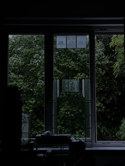 Blick durch ein, drei geteiltes Fenster in den abendlichen Garten. Ein großer Baum nimmt den gesamten Hintergrund ein. Im oberen Teil des Fensters hängen drei markierte kleine Bleiglas Fensterscheiben. Rings im Vordergrund, die hohe Box eines Verstärker.