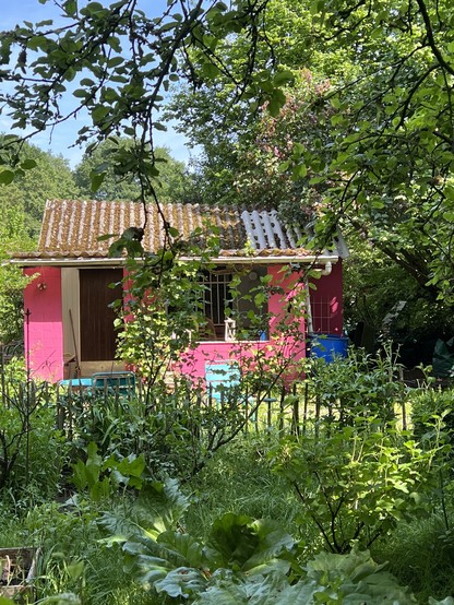 A small, brightly red colored house with a weathered roof sits in a lush garden surrounded by greenery and trees 

Ein kleines, leuchtend rot gefärbtes Haus mit einem verwitterten Dach befindet sich in einem üppigen Garten, der von Grün und Bäumen umgeben ist.
