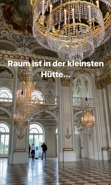 A lavish baroque-style room with ornate chandeliers, high ceilings, decorative molding, arched windows, and marble flooring. Two people are seen in the background near the windows. The text in German reads, 