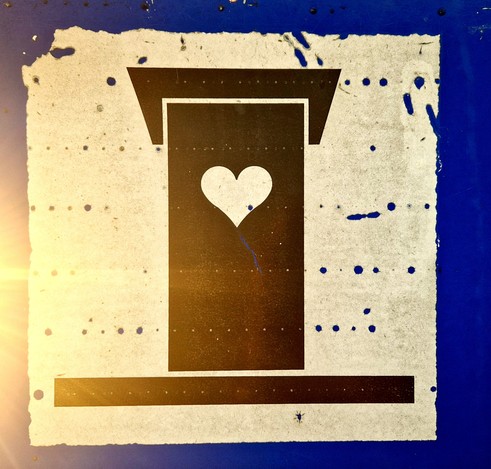 Weisses Schild mit Toilettenhaus und großem Herz auf blauem Hintergrund.