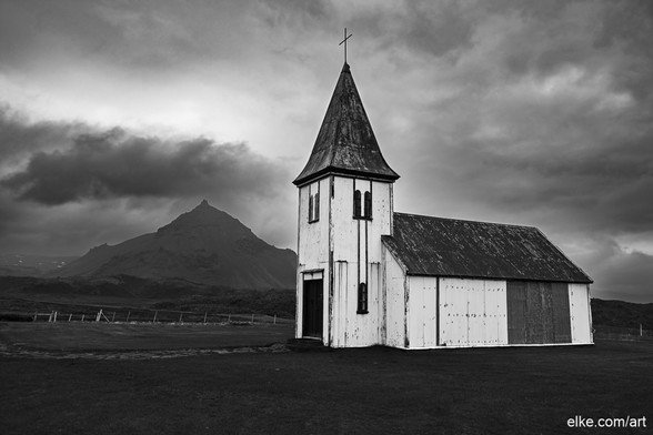 This striking black and white photograph captures Hellnarkirkja, a quaint wooden church set against the dramatic backdrop of a looming mountain and ominous clouds in Hellnar, Iceland.

The church's weathered exterior and sharp steeple provide a stark contrast to the rugged natural landscape, evoking a sense of isolation and solemn beauty. The composition, with the mountain peak partially shrouded in mist, adds an ethereal quality to the scene, making it a poignant piece that highlights the intersection of human architecture and untouched nature.