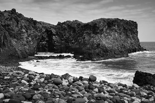 This black and white photograph captures the rugged beauty of the Hellnar coastal cliffs in Iceland.

The striking geological formations, with their jagged edges and dark basalt rock, stand in stark contrast to the turbulent waves crashing below. The rocky shore is covered with smooth, rounded stones, adding texture to the scene. The dramatic landscape evokes a sense of timelessness and raw natural power, making it a captivating piece for any art collection.