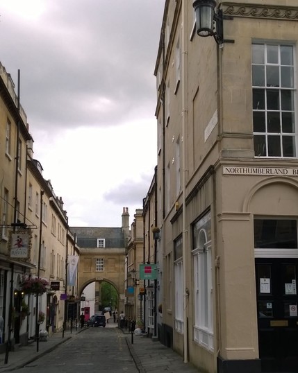 A narrow, cobblestone street lined with historic sandstone buildings, featuring signage for businesses including 