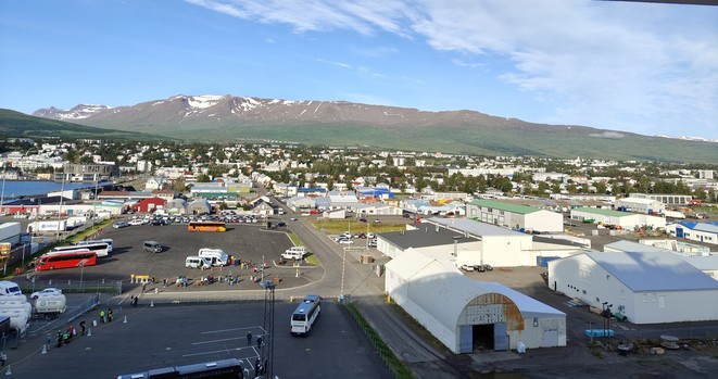 Panorama von Akureyri vom Schiff aus gesehen. Im Vordergrund befinden sich die Hafenanlagen und Hallen. Dahinter erstreckt sich die Stadt, ausschließlich bestehend aus ein- oder zweistöckigen Gebäuden. Hochhäuser gibt es keine. Im Hintergrund erkennt man hohe Bergrücken, teilweise vereist. Der Himmel ist blau, teilweise gibt es ein paar Wolken.