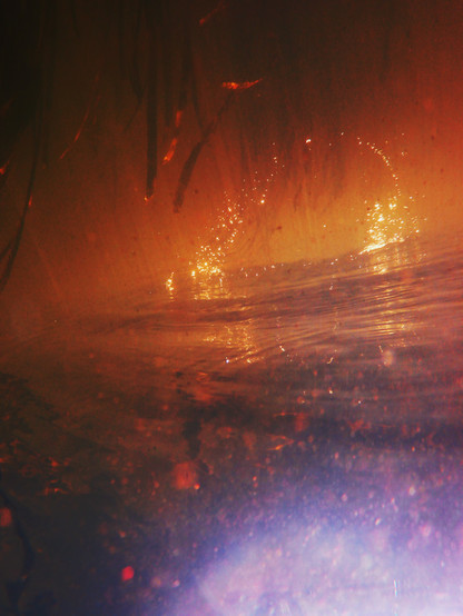 Das Foto zeigt eine abstrakt verfremdete Unterwasser-Aufnahme. Unten gibt es blautöne, darüber dominieren rot-orangene Töne. Man sieht einen glitzernden Bogen, der auf einer Wasseroberfläche zu stehen scheint. Es wirt sehr fremdartig.