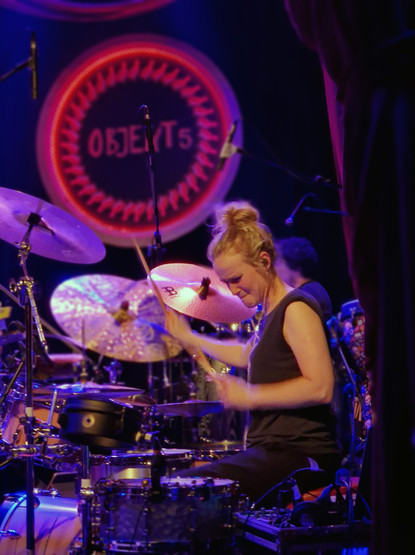 Anika spielt gerade Schlagzeug. Sie saß längs zum Bühnenrand, so dass sie von der Seite zu sehen ist. Im Hintergrund ist das grosse Logo 