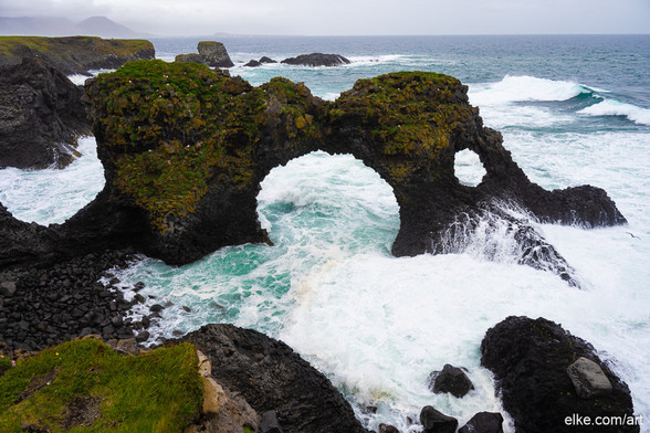 This stunning image captures the breathtaking beauty of Gatklettur, a natural rock arch located near Arnarstapi on the coast of Iceland.

The rugged basalt formations, covered in patches of green moss, rise dramatically from the churning, turquoise waters of the Atlantic Ocean. The arch, shaped by centuries of relentless waves, stands as a testament to the raw power and beauty of nature. The surrounding cliffs and distant rocky islets shrouded in mist and the ever-changing weather of Iceland, create a moody, almost otherworldly atmosphere.

The vibrant contrast between the dark volcanic rock, the vivid greenery, and the swirling white sea foam add depth and dynamism to the scene.