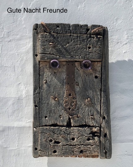 A piece of weathered wood resembling a face, mounted on a white wall, with the text 