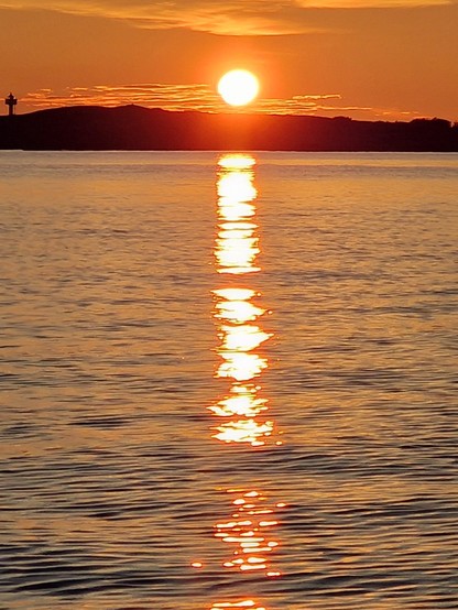 Sonnenuntergang über Stensholem. Links auf der Insel steht ein kleines Seezeichen.  Die Sonne ist noch deutlich am Himmel zu sehen, sie spiegelt sich ber schon rötlich im Wasser. Sonne und Spiegelung sehen Munch-typisch wie ein 