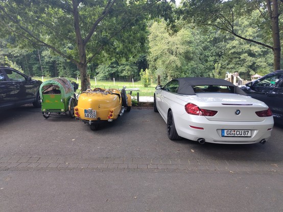 Links ein grünes Lastenrad, etwa 80 cm breit, daneben ein historisches Cabriolet mit etwas 120 cm Breite zuletzt ganz rechts ein moderner BMW,