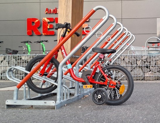 Rotes Kinderfahrrad mit Stützrädern steht allein in großem Fahrradständer, dessen Bügel höher hinaus ragen als das zierliche Rädchen groß ist.