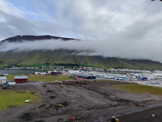 Sicht vom Schiff auf Ísafjörður. Im Vordergrund befindet sich eine große Brache, die gerade Baustelle ist. Dahinter erkennt man die Stadt selbst und einen Teil des Fjords. Im Hintergrund ragt ein Bergmassiv auf. Wolken bedecken teilweise das obere Drittel des Massivs.