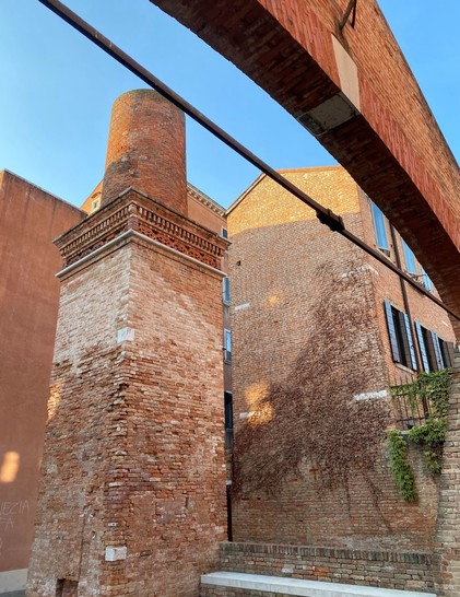 Brick buildings with architectural details under a clear blue sky. A large chimney, archway, and some climbing plants are visible.

Backsteingebäude mit architektonischen Details unter einem klaren blauen Himmel. Ein großer Schornstein, ein Torbogen und einige Kletterpflanzen sind sichtbar.