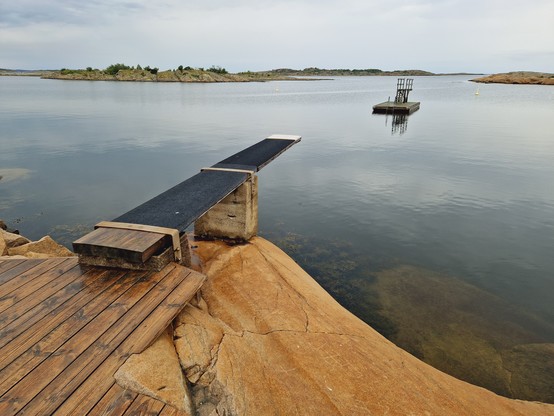 Ganz ruhige See. Von links unten ragt ein Sprungbrett über das Wasser. In der Mitte rechts schwimmt eine kleine Holzinsel mit Sprungturm im ruigen Wasser. Morgenlicht. Im Hintergrund sieht man Felseninseln.