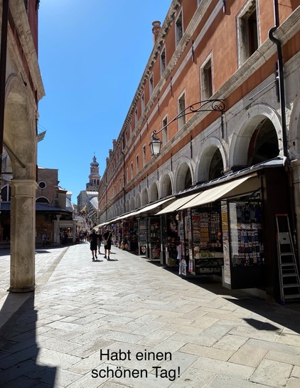 A sunny street scene featuring a narrow walkway lined with shops selling souvenirs. There are two people walking along the path, with historic buildings and a prominent tower visible in the background. A message in German reads, 