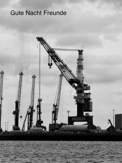 A black and white image of a large crane and several smaller cranes at a port, with a cylindrical object on the dock. The sky is overcast, and the text 