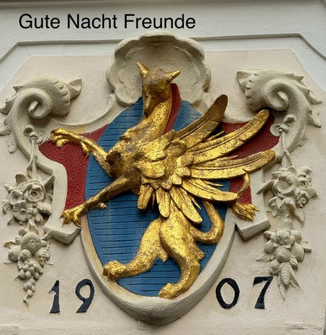 A decorative sculpture featuring a gold griffin with wings, set against a blue shield, alongside red and beige elements. The year 