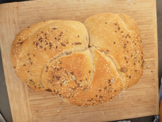 Ein Brot auf einem Holzbrett. Es ist zu erkennen, dass das Brot vor dem Backen geflochten wurde. Auf dem Brot sind mitgebackene  Körner zu sehen.