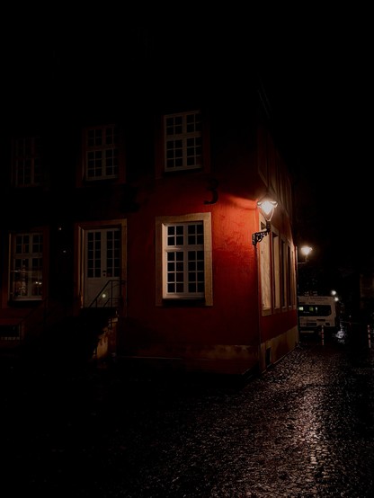 A dimly lit street at night featuring the corner of a red building. Soft light from a streetlamp illuminates part of the facade and nearby cobblestone pavement. Windows are visible, and there is a staircase leading to the entrance.

Eine matt beleuchtete Straße bei Nacht mit der Ecke eines roten Gebäudes. Weiches Licht einer Straßenlaterne beleuchtet einen Teil der Fassade und den nahe gelegenen Kopfsteinpflasterpflaster. Fenster sind sichtbar, und es gibt eine Treppe, die zum Eingang führt.