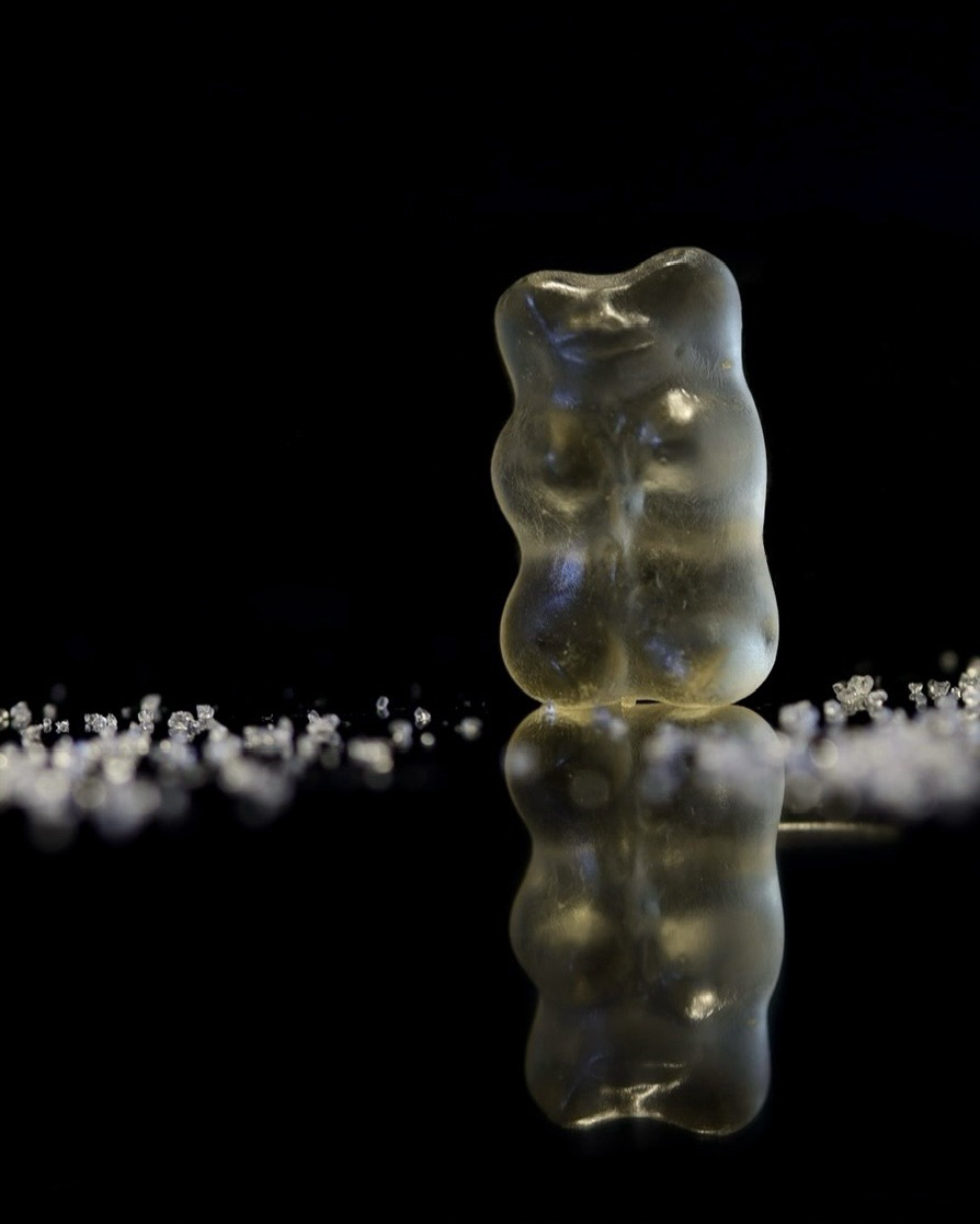 Makroaufnahme: Ein helles Gummibärchen vor schwarzem Hintergrund und auf schwarzer spiegelnder Fläche, daneben einige weiße Zuckerkristalle, alles komplett gespiegelt.