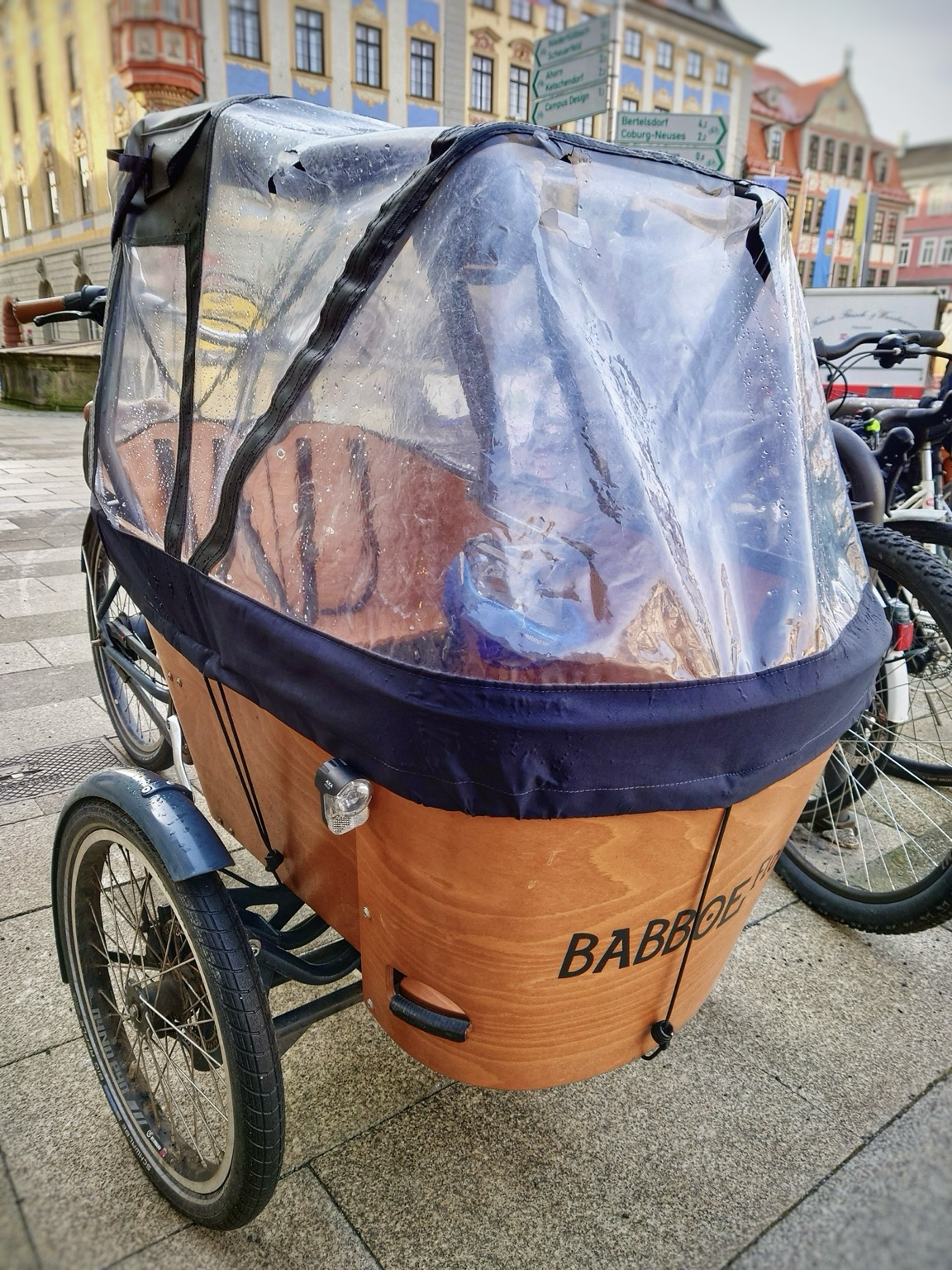 Ein Lastenfahrrad von vorne rechts, die helle Holzwanne zwischen den Vorderrädern hat einen transparenten Aufbau mit Plastik-Regenschutz. Links dahinter ist das Hinterrad zu erkennen. Im Hintergrund unscharf historische Gebäude.