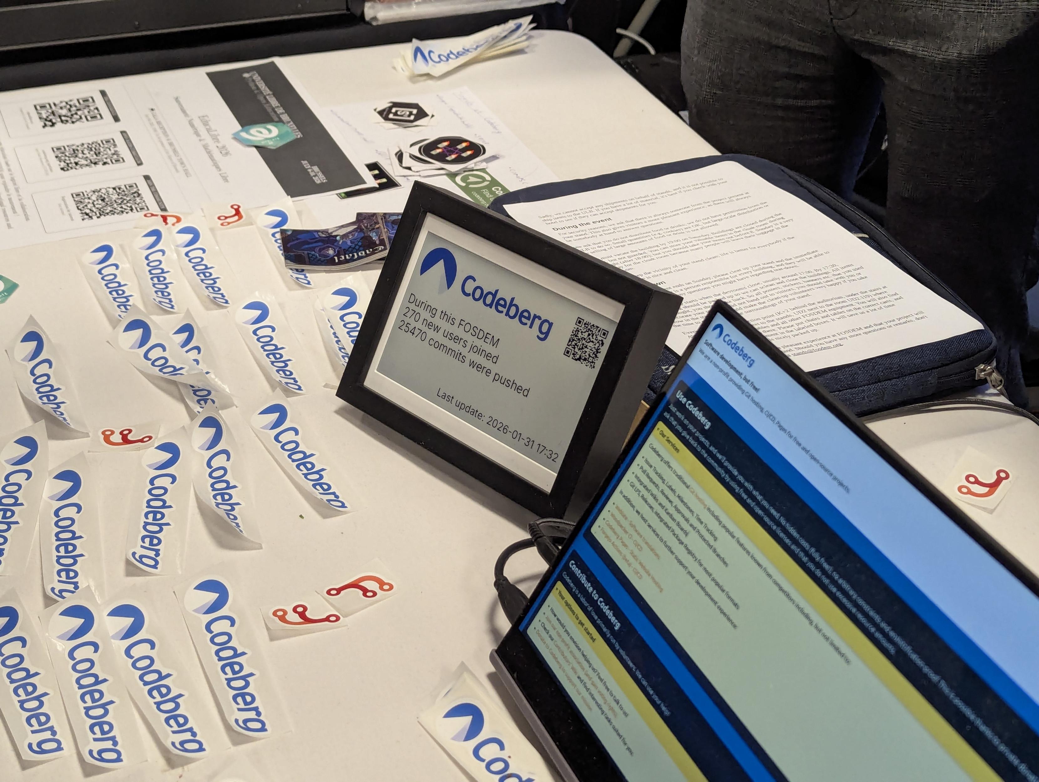 e-Paper depicting Codeberg's stats "since the beginning of FOSDEM".
During this FOSDEM, 270 new users joined. 25740 commits were pushed.
There's a display depicting stand.codeberg.org to the right, and many visible Forgejo and Codeberg stickers to the left.