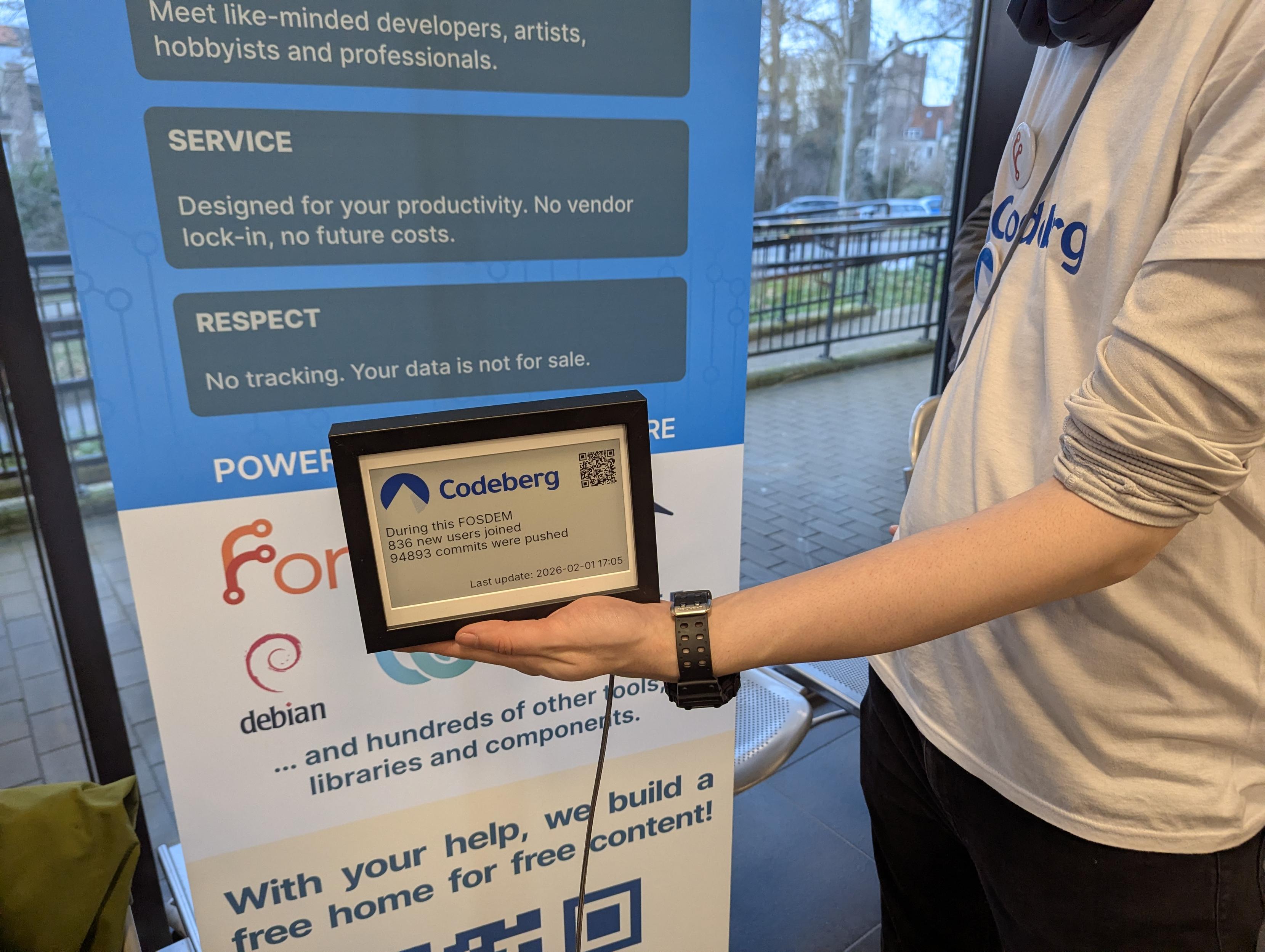 Person with a Codeberg T-shirt and a watch holding the Codeberg e-paper display in front of a roll-up of Codeberg. The e-paper sign displays the following stats are present. "During this FOSDEM, 836 users joined, 94893 commits were pushed. Last updated today, 17:05 CET.