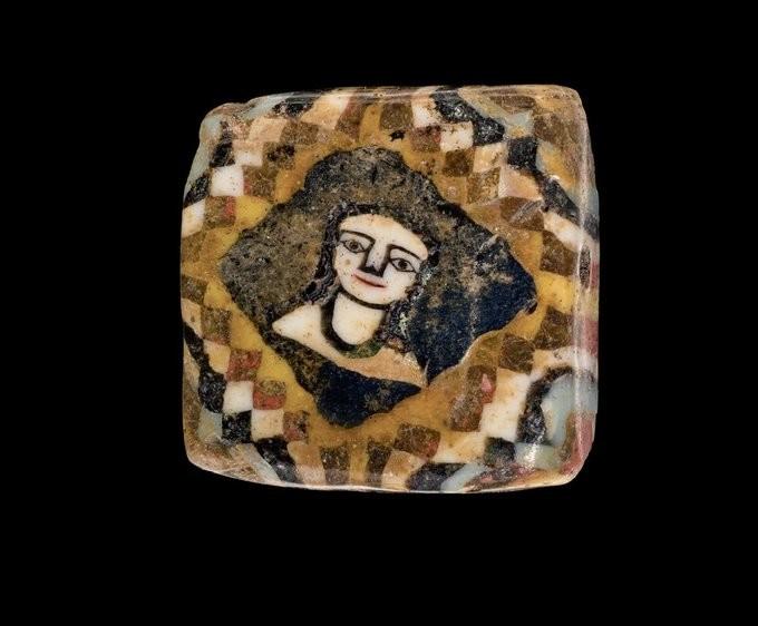 Square glass bead featuring a checkerboard mosaic border in tan, white, black, and reddish tones, surrounding a female face with prominent white eyes and a subtle smile on a black background