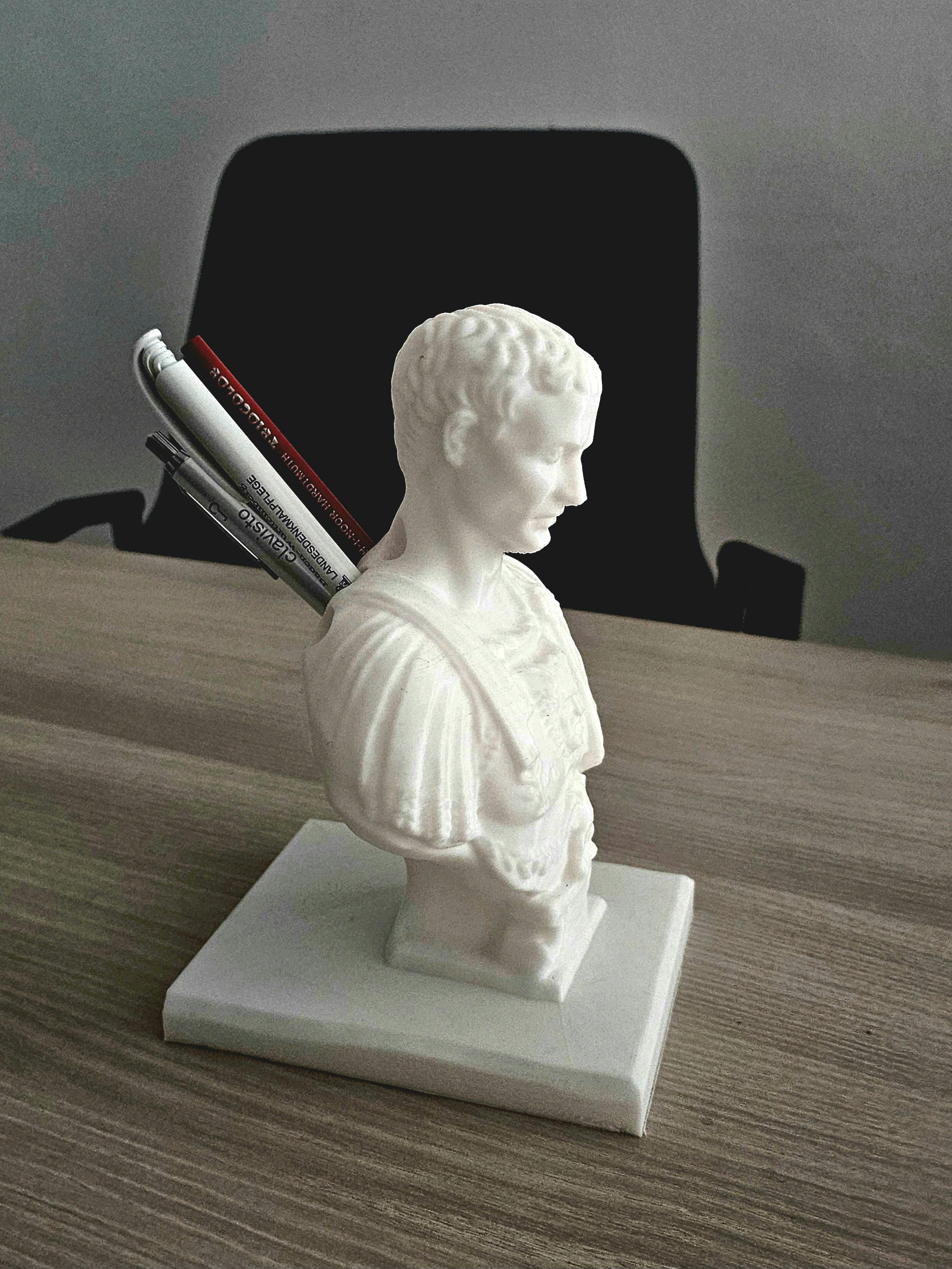 A white pen holder shaped like a bust of Julius Caesar sits on a light wooden desk. Several pens and pencils are inserted into the back of the bust, humorously resembling knives. A large black office chair and a blurred white wall are visible in the background.