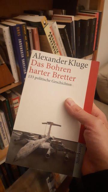 Eine Hand hält das Buch "Das Bohren harter Bretter" von Alexander Kluge vor einem Buchregal. Im Hintergrund unter anderem die Bücher "Sand" von Wolfgang Herrndorf und "Absolute FreeBSD".