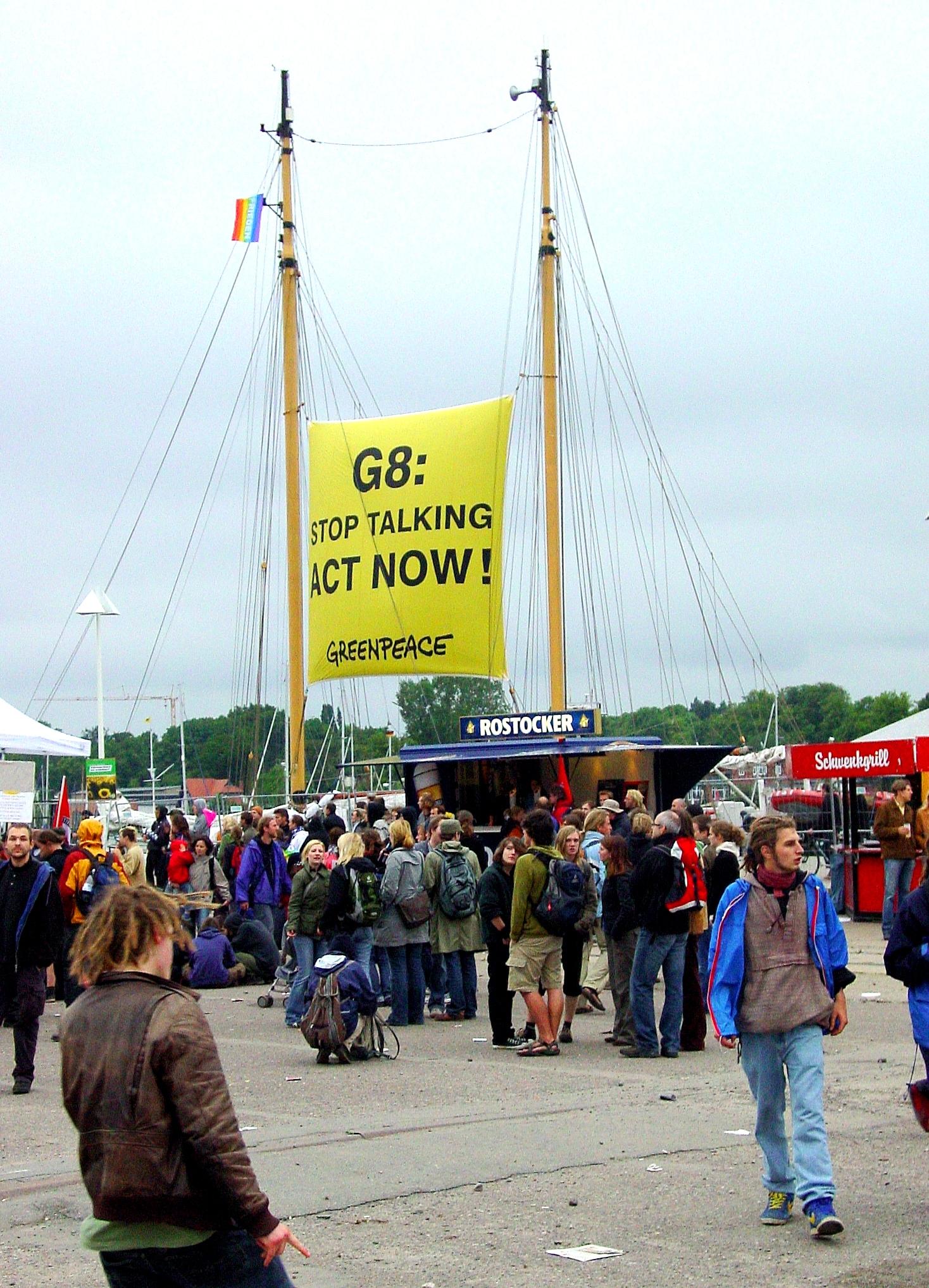 Man sieht die 2 Masten eines Segschiffes. Dazwischen hängt ein Transparent "G8: Stop Talking ... ACT now Greenpeace"