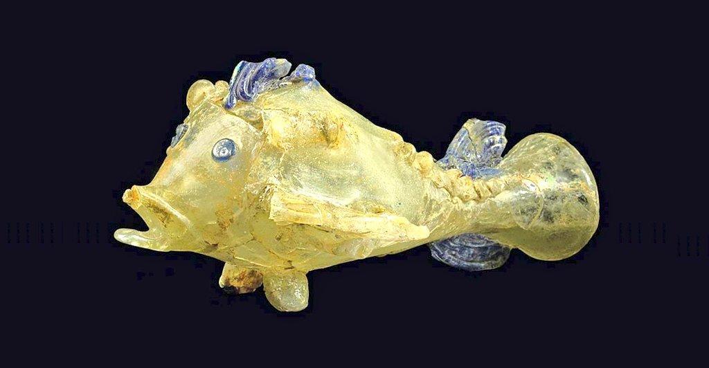 Fish-shaped glass vessel with translucent yellowish body, wide open mouth, round eyes, and blue accents on fins, crest, and tail