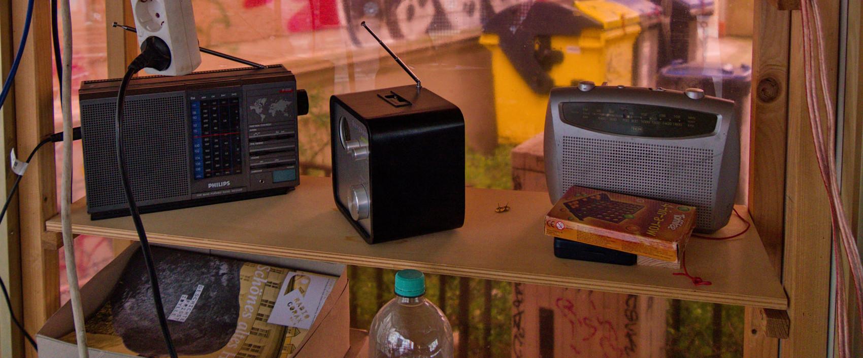 Drei verschiedene Radios auf einem Regal. Ein Brett tiefer ist ein Stapel Programmzeitungen von Radio Corax zu sehen.