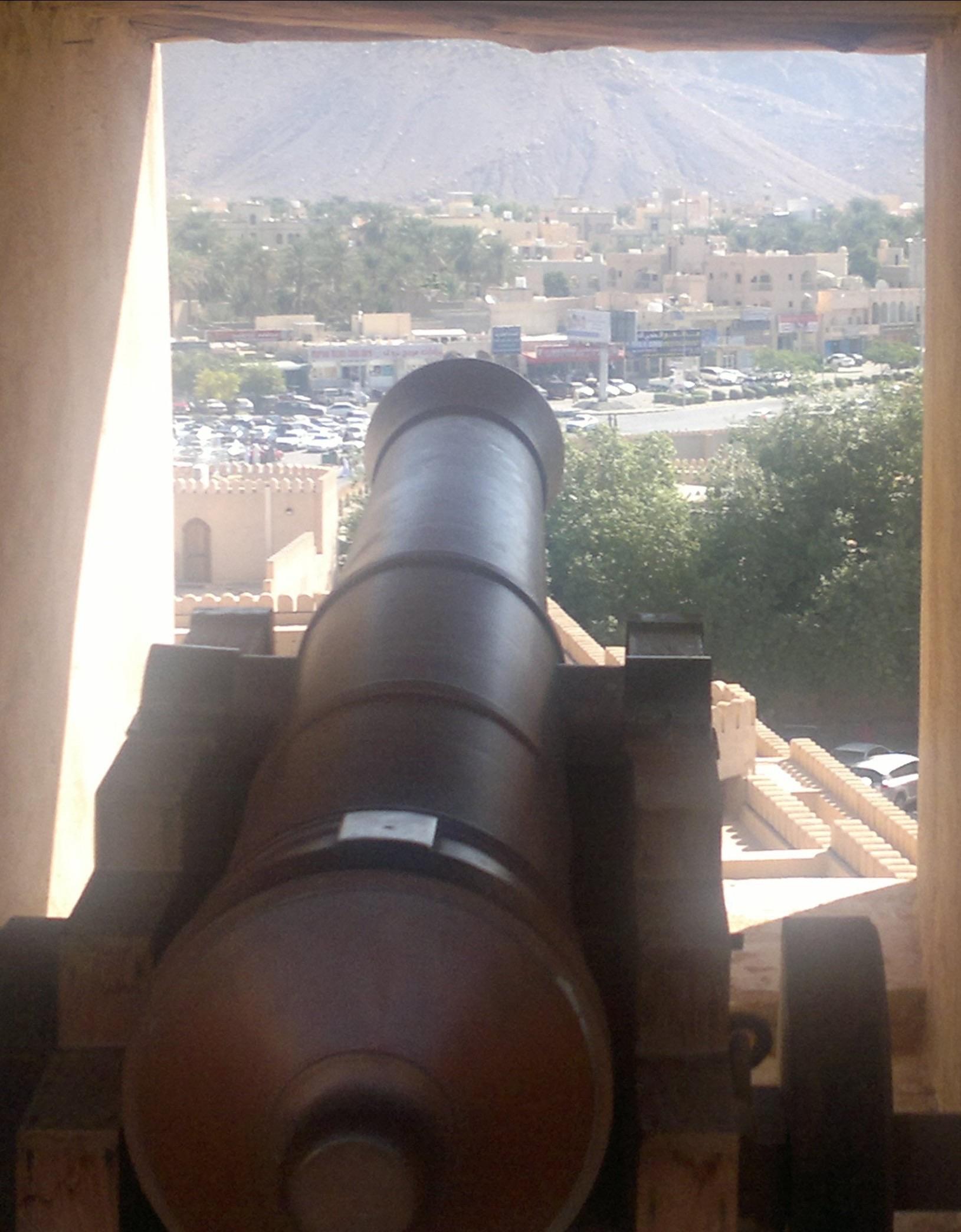 A historic cannon is positioned at a lookout, framed by a stone wall. In the background, a view reveals a modern town with buildings, palm trees, and parked cars. The scene contrasts the ancient cannon with contemporary architecture.
Auf einem Aussichtspunkt steht eine historische Kanone, eingerahmt von einer Steinmauer. Im Hintergrund bietet sich der Blick auf eine moderne Stadt mit Gebäuden, Palmen und geparkten Autos. Die Szene stellt die alte Kanone in Kontrast zur zeitgenössischen Architektur.