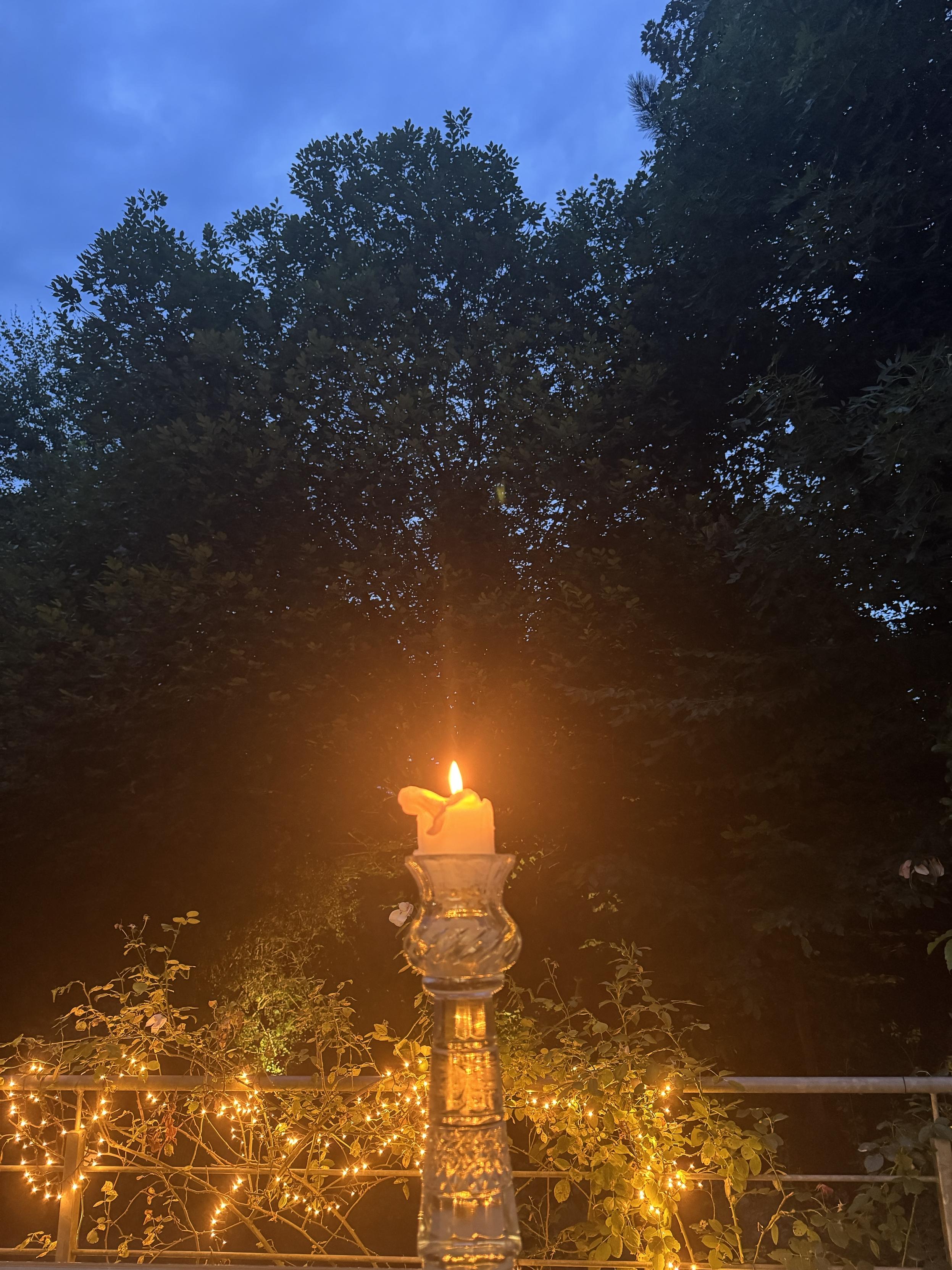 A lit candle in a glass holder stands in the foreground, illuminating a dimly lit outdoor space adorned with twinkling lights. In the background, tall trees and a cloudy sky create a serene, moody atmosphere.
Im Vordergrund steht eine brennende Kerze in einem Glashalter, die einen schwach beleuchteten Außenbereich erhellt, der mit funkelnden Lichtern geschmückt ist. Im Hintergrund sorgen hohe Bäume und ein bewölkter Himmel für eine ruhige, stimmungsvolle Atmosphäre.