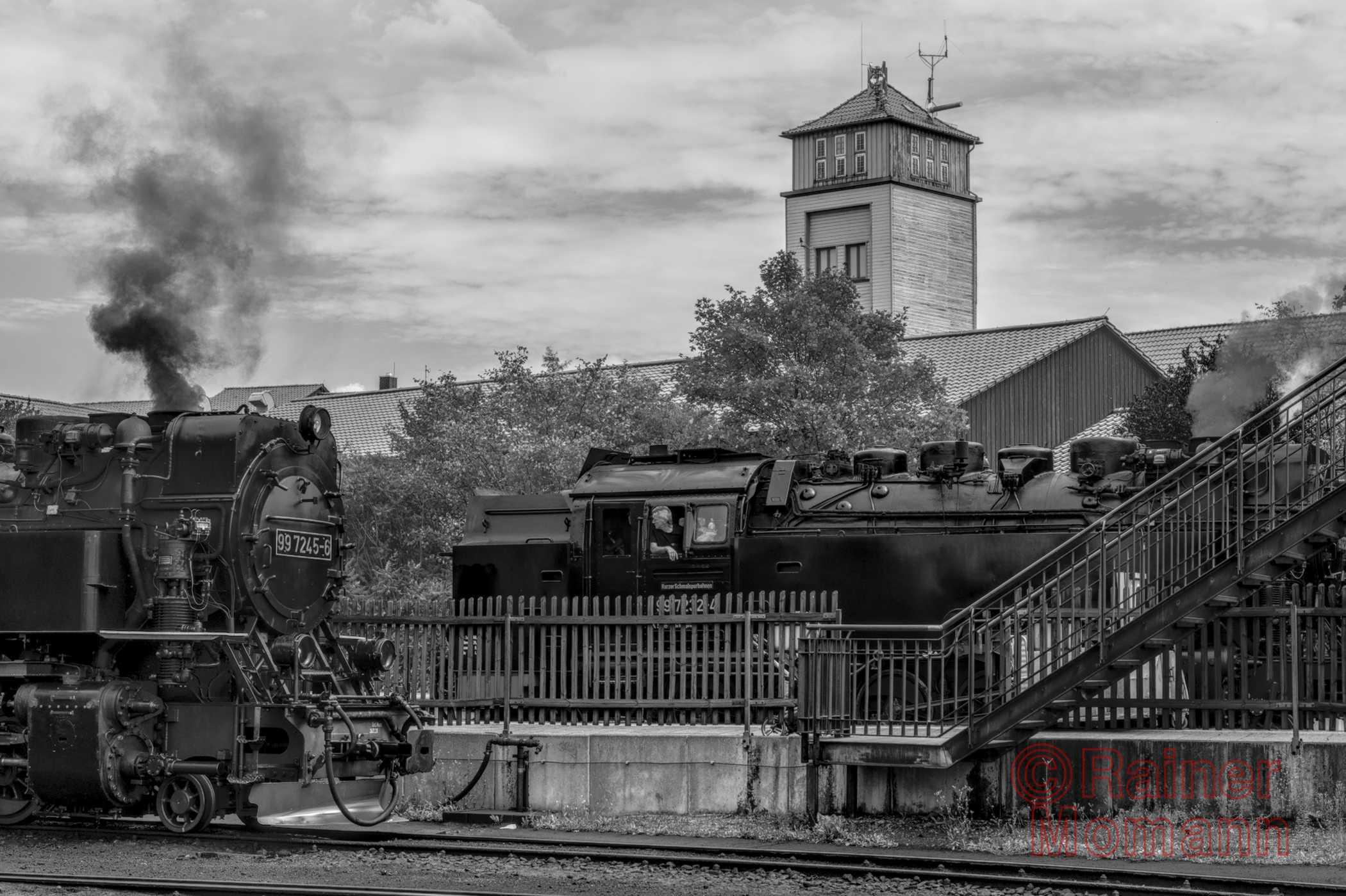 Schwarzweißfotografie im Format 3:2 Landscape.
Auf einem Bahnhof stehen zwei Dampflokomotiven auf unterschiedlichen Gleisen. Im Hintergrund Bäume, hinter welchen ein Gebäude mit einem hohen quadratischen Turm hervorschaut.