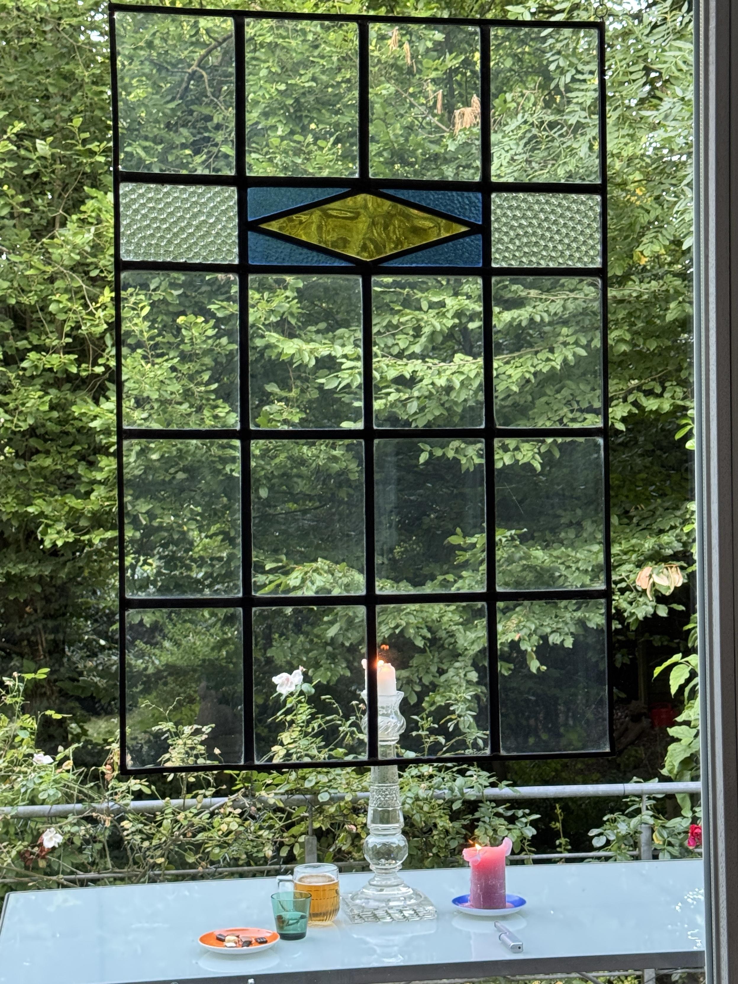 Blick durch eine Bleiverglasung in den grünen Garten. Auf dem Terrassentisch brennen Kerzen eine davon in einem riesigen Kerzenleuchter. Man sieht noch ein Glas mit Bier, ein kleines grünes Trinkglas und eine Untertasse