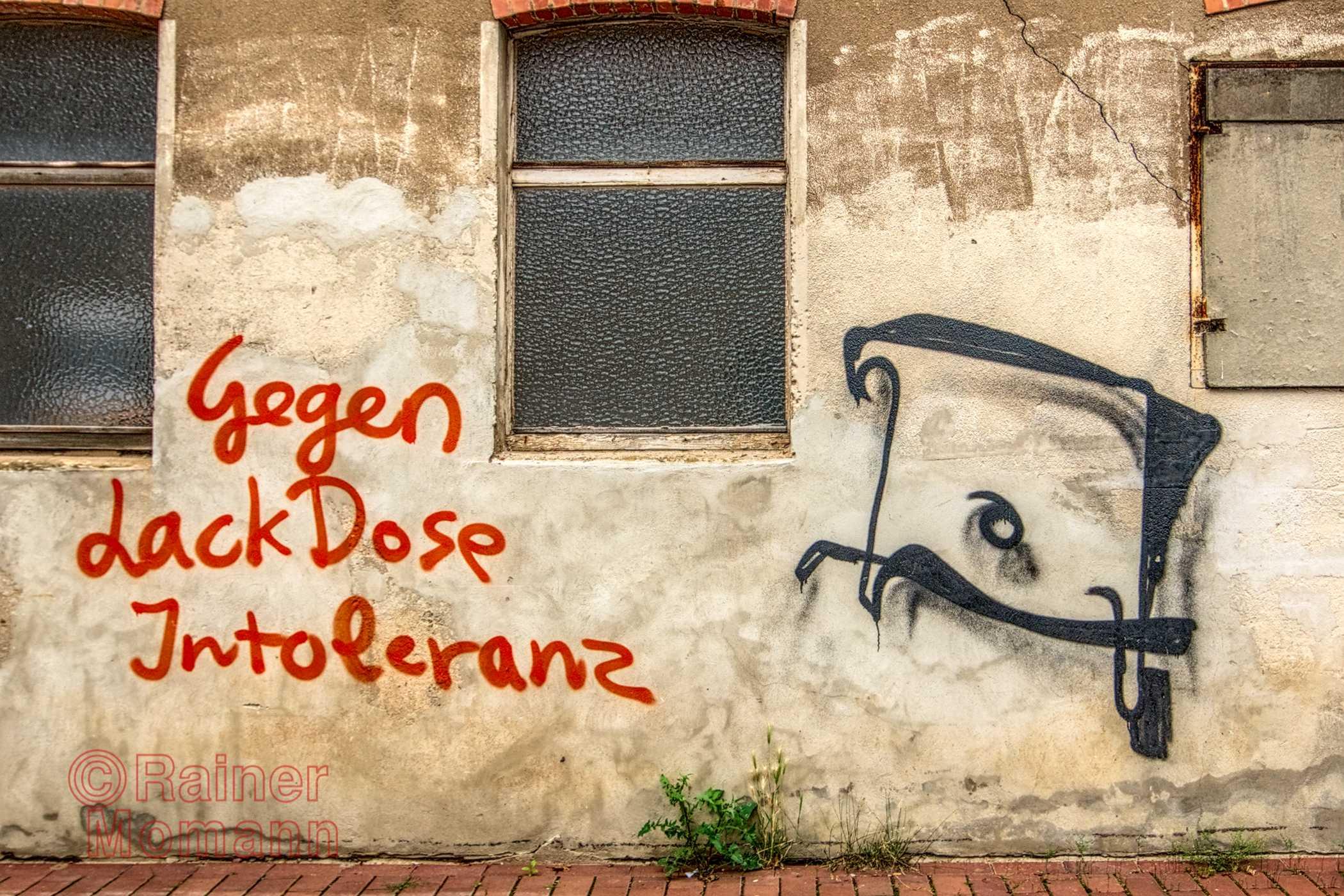 Farbfotografie im Format 3:2 Landscape.
Auf einer Wand mit altem, verschmutzen Putz und drei Fenstern, steht in roter Farbe der Spruch 'Gegen LackDose Intoleranz'. Daneben in Schwarz, ein einfaches, rautenförmiges Graffiti.