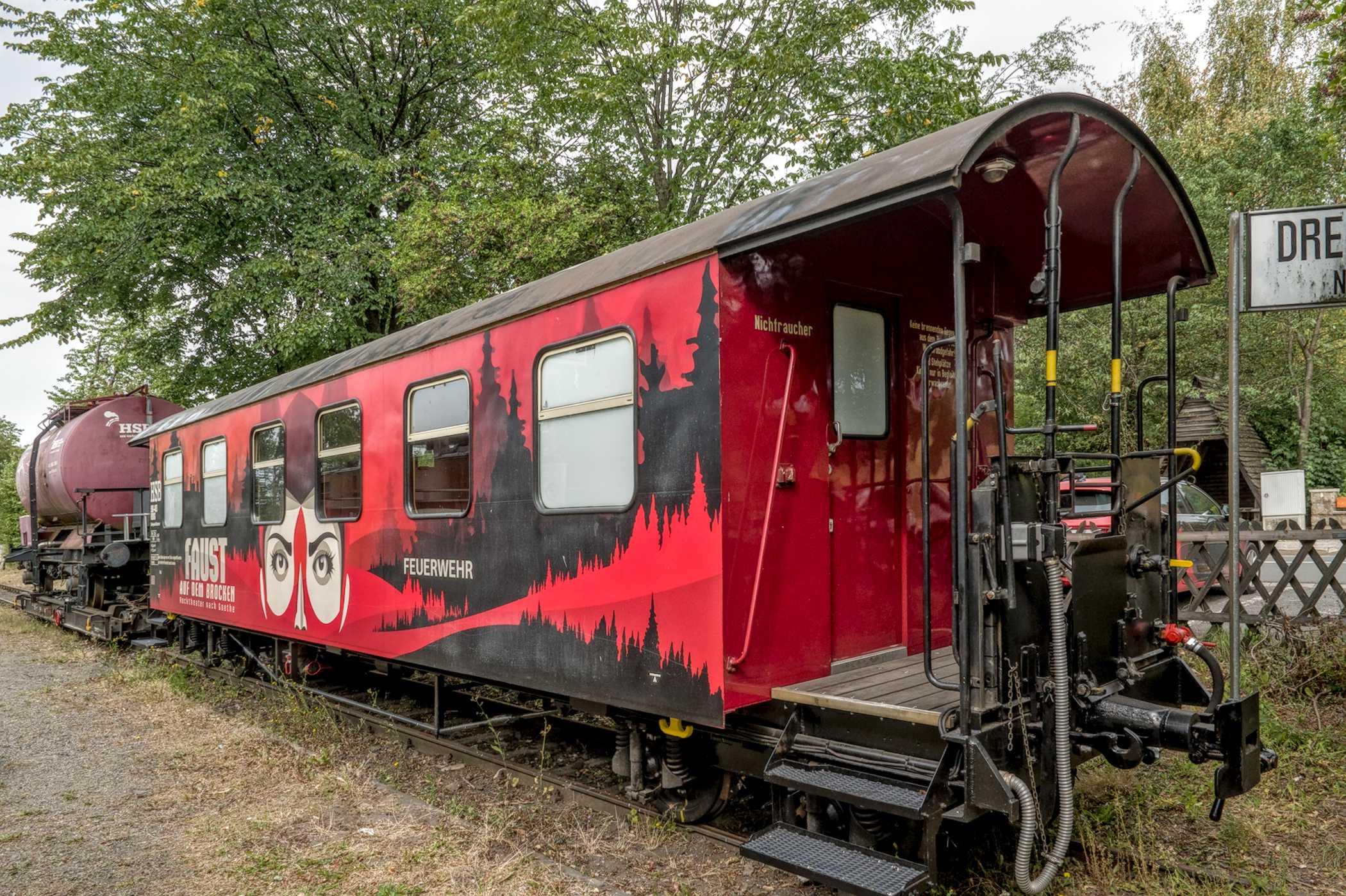 Farbfotografie im Format 3:2 Landscape.
Ein alter Personenwagon der Harzer Schmalspurbahn. Er ist mit einem in rot-schwarz gehaltener Harzer Landschaft, sowie mit der weißen Schrift "FEUERWEHR" bemalt. Des Weiteren ist ein Gesicht in schwarz-weiß zu sehen.