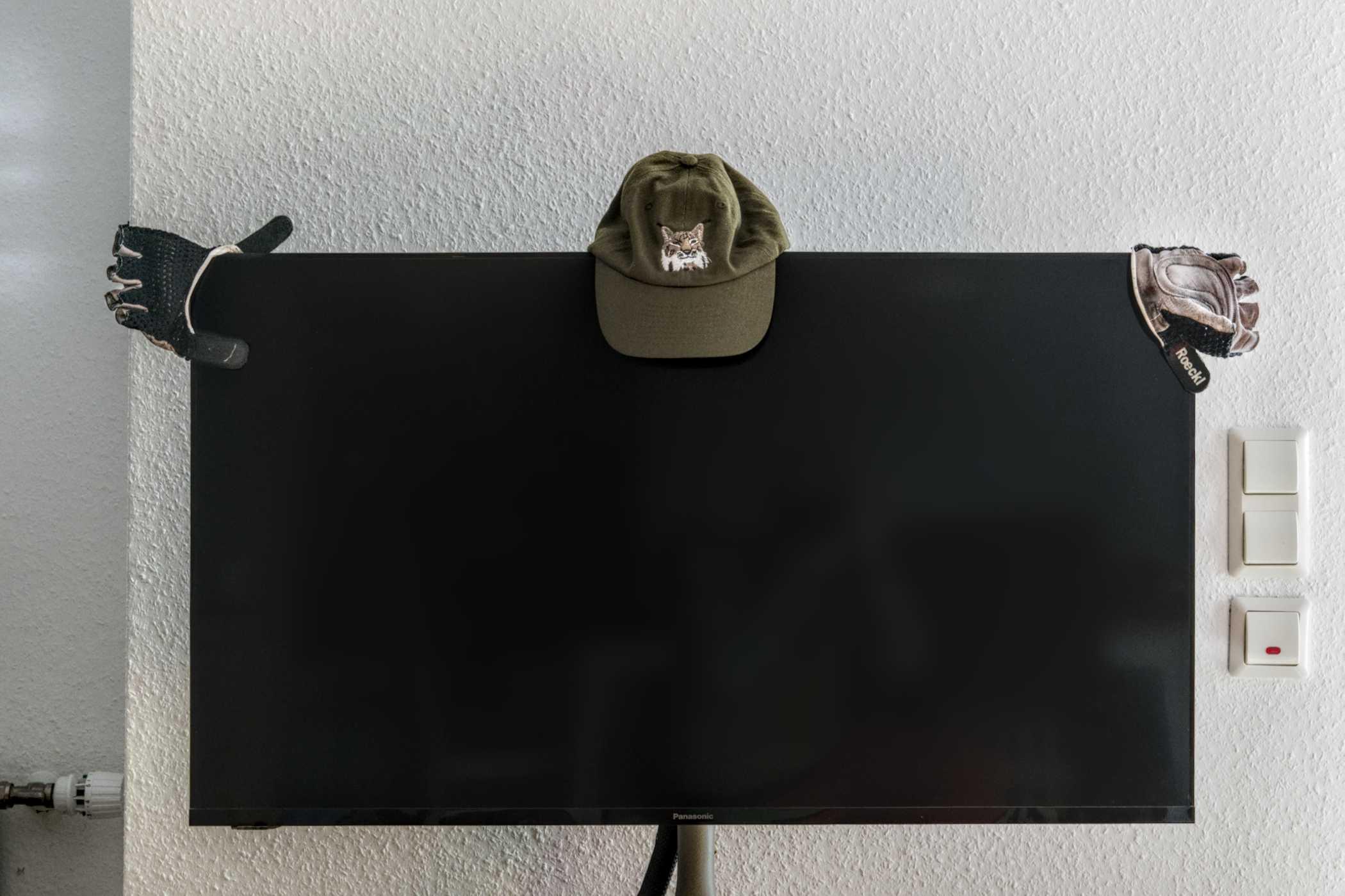 Ein ausgeschalteter Flachbildfernseher vor einer weißen Wand. Auf dem oberen Rand des Gerätes sind in der Mitte eine grüne Basecap, sowie rechts und links auf den oberen Ecken, jeweils ein Fahrradhandschuh aufgesetzt.