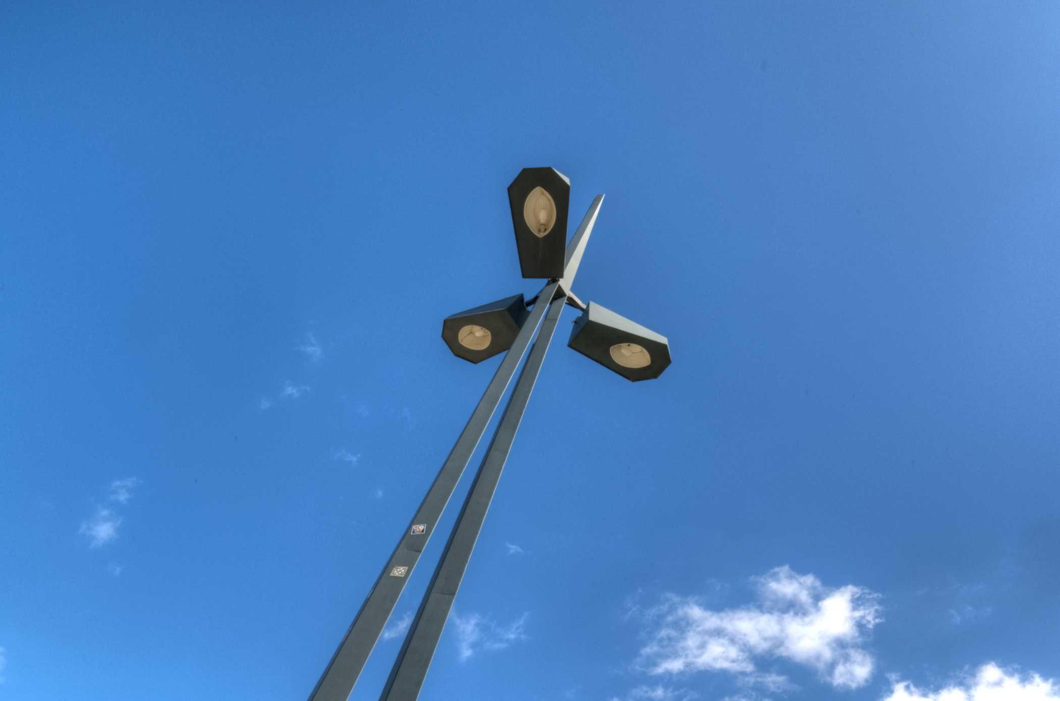 Farbfotografie im Format 3:2 Landscape.
Blauer Himmel mit wenigen weißen Kumuluswolken am unteren Bildrand. Von unten, links der Bildmitte, nach rechts oben, ein zweigeteilter Laternenmast, dessen rautenförmige Körper oben in einer schmalen, langen Spitze vereint sind. Unterhalb der Spitze sind drei tropfenförmige Lampenkörper, symmetrisch angebracht.