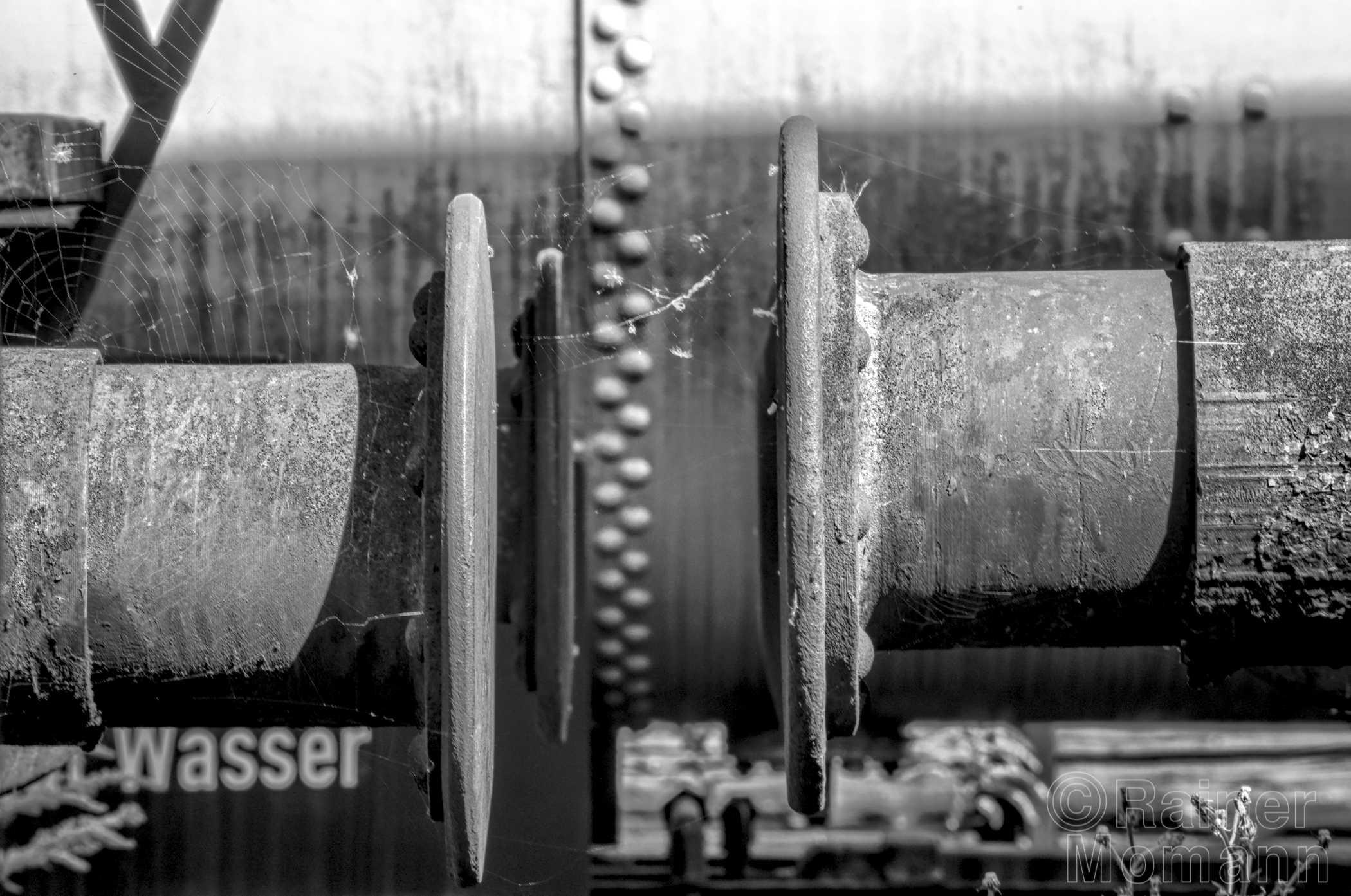 Schwarzweißfotografie im Format 3:2 Landscape.
Zwei Pufferpaare alter Eisenbahn Wagons, mit etwas Abstand und in leicht unterschiedlicher Hohe. Im Hintergrund, etwas unscharf, ein runder Tankwagon welcher rundherum mit einer Doppelreihe runder Nietenköpfe.