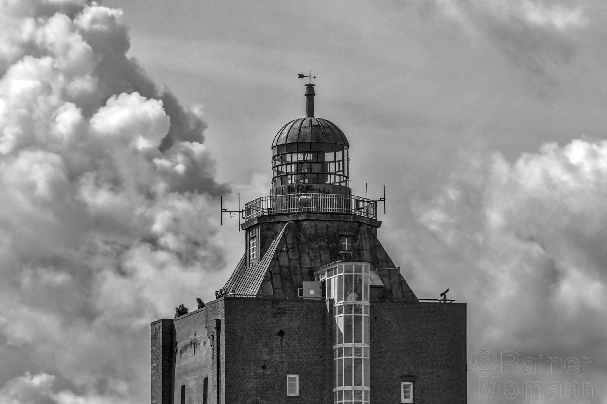 Schwarzweißfotografie im Format 3:2 Landscape.
Vor einem mit kräftigen Wolken durchzogenen Himmel, ist der obere Teil des quadratischen, aus Ziegelsteinen errichteten Leuchtturms von Neuwerk zu sehen.