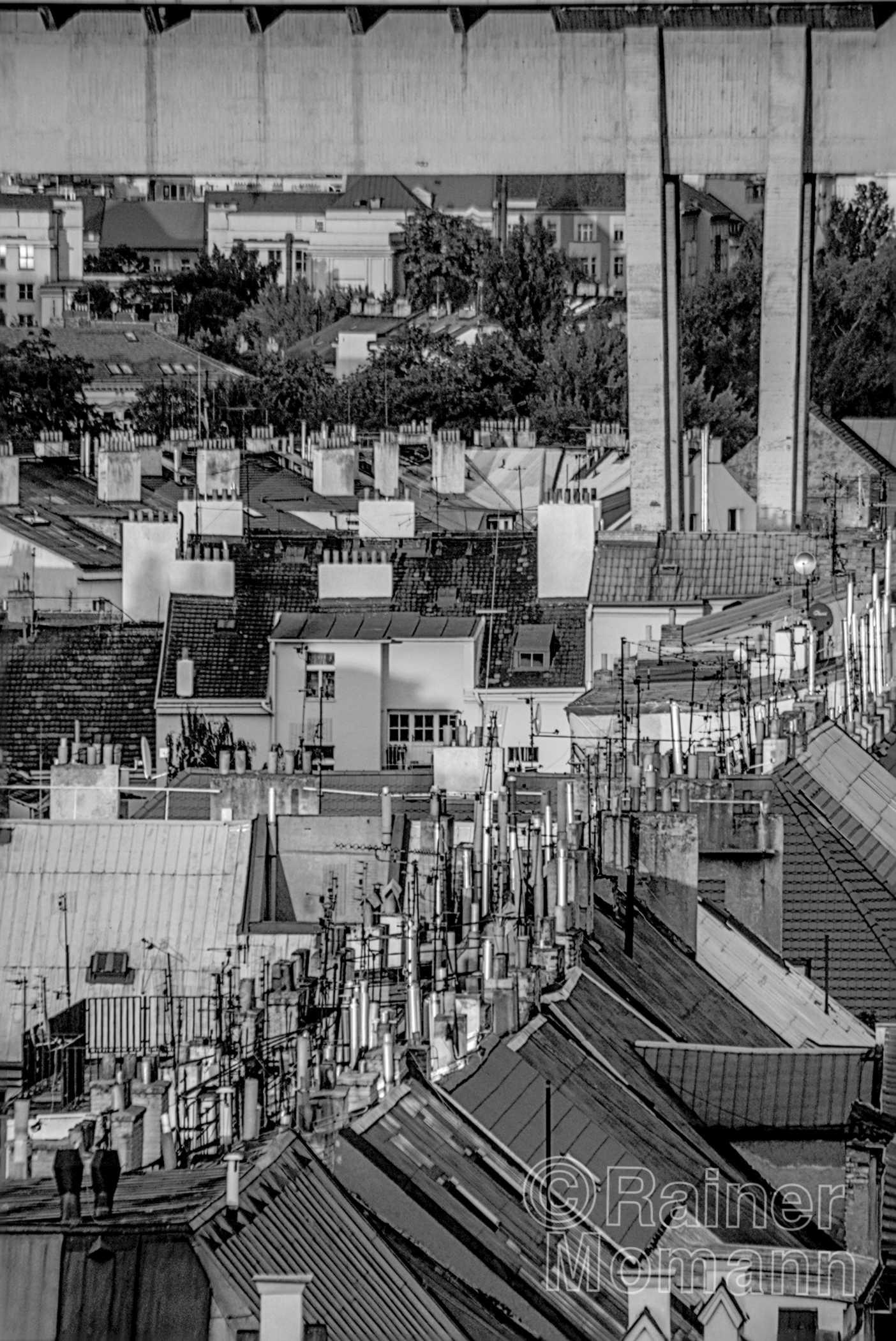 Schwarzweißfotografie im Format 2:3 Portrait.
Viele Giebeldächer mit unterschiedlichster Deckung und mit noch viel mehr Kaminen und Entlüftungsrohren. Dazwischen unterschiedlichste Antennen und am gesamten oberen Bildrand, anstatt Himmel, ein langer grauer Kasten welcher rechts von zwei dünnen, kantigen, zwischen den Dächern verschwinden Stützen verschwinden.